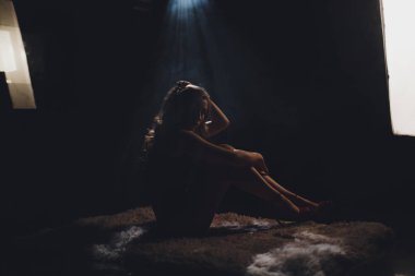 Stüdyo ışık altında oturan, dizlerinin üzerine eğilerek ve kamera darck özgeçmişlerine bakarak güzel genç kadın silüeti.