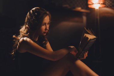 Uzun kahverengi saçları siyah seksi iç çamaşırı bookshelf bir kitapta seyir altında oturan şehvetli genç kadınla floot lamba tarafından karanlık bir arka plan üzerinde yaktı..