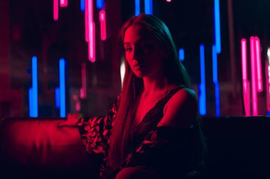 Deri kanepede oturan şehvetli genç kadın kırmızı neon ışık ve neon tüplerin bir arka planda bir duvar ile yaktı