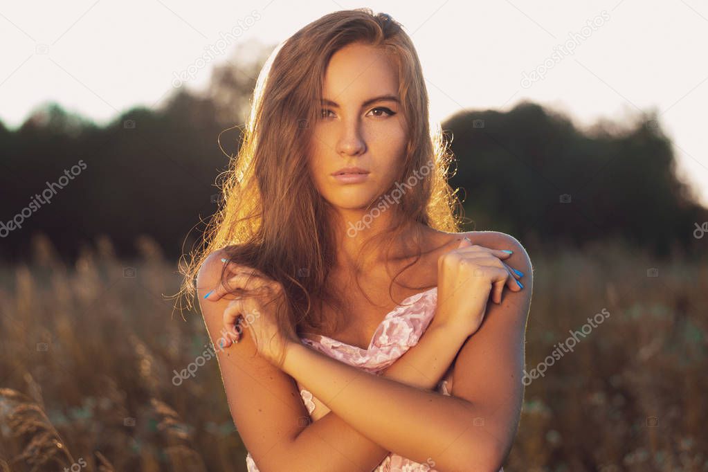 Cute young woman in silk transparent scarf standing looking in camera back lit by sunset light