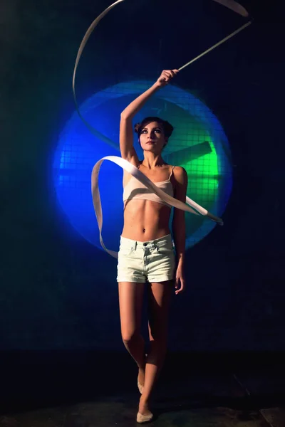 Young gymnast woman training with a gymnastics tape against the black wall with a big fan lit with blue and green neon light