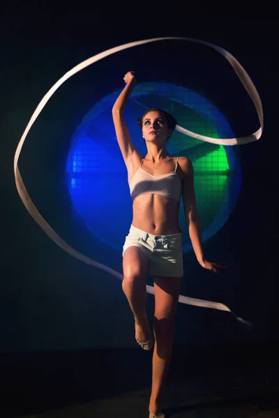 Beautiful young gymnast woman training with a gymnastics tape against the black wall with a big fan lit with blue and green neon light