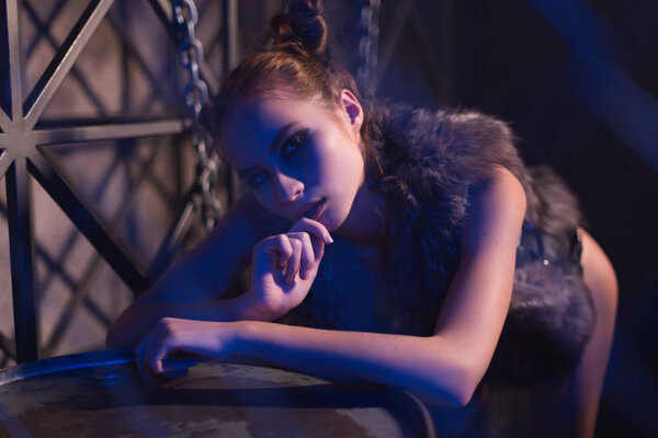 Sensual young woman posing topless or nude in fur jacket in steampunk style scene leaning on metal barrel on a dark moody background.