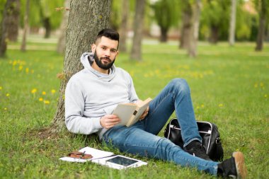Genç adam çimlerin üzerinde oturuyor ve tablet kullanıyor.