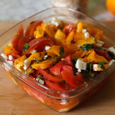 Dilimlenmiş domatesli taze bir salata, sarı dolma biber ve cam kasede parçalanmış peynir. Salata otlarla süslenir ve ahşap bir yüzeyde servis edilir.