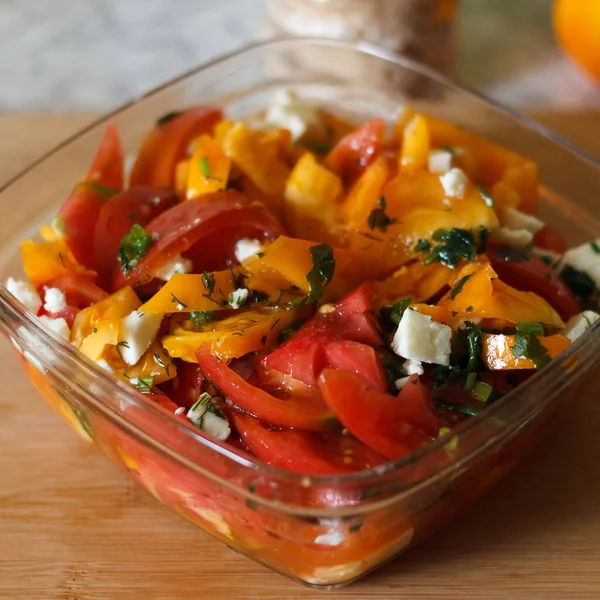 Dilimlenmiş domatesli taze bir salata, sarı dolma biber ve cam kasede parçalanmış peynir. Salata otlarla süslenir ve ahşap bir yüzeyde servis edilir.