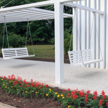 Minimalist bir yapının altında beyaz salıncakların olduğu modern bir açık hava alanı. Bölge yeşillik ve çiçek yataklarıyla çevrili, dingin bir ortam yaratıyor.