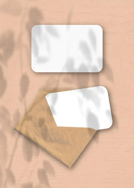 Sebuah amplop dengan dua lembar kertas putih bertekstur pada latar belakang persik meja. Mockup overlay dengan bayangan tanaman. Cahaya alami melemparkan bayangan dari tanaman eksotis — Stok Foto