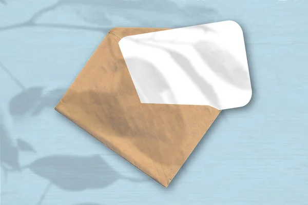 Ein Umschlag mit einem Blatt strukturiertem weißem Papier auf blauem Hintergrund. Mockup mit einer Überlagerung von Pflanzenschatten. Natürliches Licht wirft Schatten von einer exotischen Pflanze. Horizontale Orientierung — Stockfoto