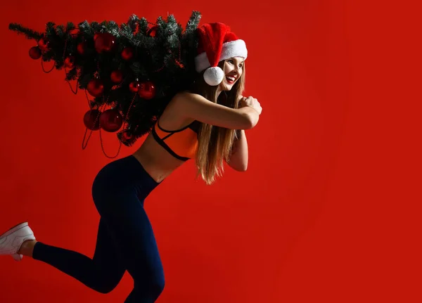 Xmas ağacı onun omuzlarında tutan Santa şapka Noel spor kadın. Kazanan enerji o kırmızı arka plan.
