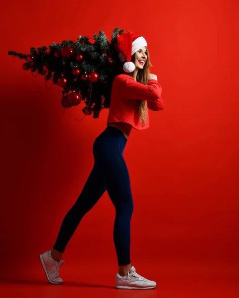 Bir Noel ağacı taşıyan bir Noel Baba şapkalı güzel bir genç atletik kız omuzunda topları ile dekore edilmiş.