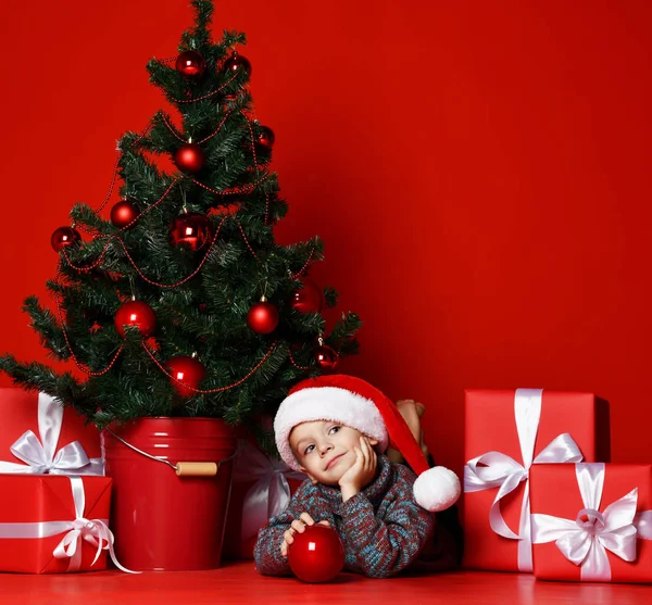 Engraçado sorrindo criança em Santa chapéu vermelho deitado no fundo da árvore de Natal — Fotografia de Stock