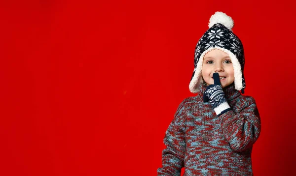 Junge mit Strickmütze und Pullover und Fäustlingen macht Schweigegegeste vor buntem roten Hintergrund. — Stockfoto