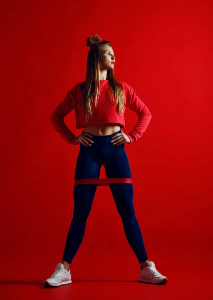 ゴムバンド付けストレッチの仕事良い体格の女性 赤の背景にファッショナブルなスポーツウェアのスポーツ少女 強度とモチベーション — ストック写真
