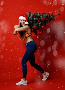 Seksi Noel fitness spor kadın Xmas ağacı onun omuzlarında tutan santa şapka giyiyor. kar taneleri