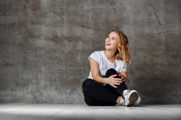 Beautiful smiling woman sitting on floor against concrete wall — Stock Photo, Image