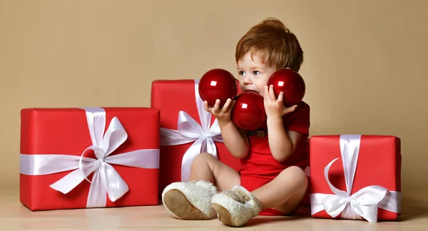 Portrait of a baby dressed a red bodysuit with big red balls decorations on the xmas present gifts over beige nude studio background. — Stock Photo, Image