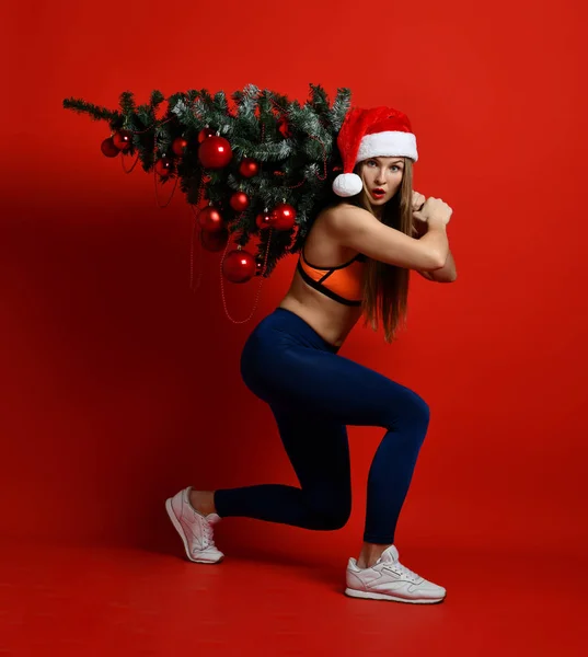 Seksi Noel fitness spor Xmas ağacı onun omuzlarında tutan Noel Baba şapkası giyen kadın.