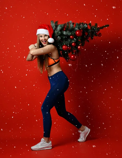 Seksi Noel fitness spor kadın Xmas ağacı onun omuzlarında tutan santa şapka giyiyor. kar taneleri