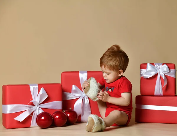 Little cute toddler boy sits among gifts dressed in a red body suit and warm sneakers. — Stock Photo, Image