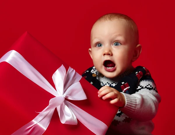 Хлопчик з великими блакитними очима в теплий светр, сидить перед його подарунком у загорнутій коробці зі стрічкою на червоному тлі . — стокове фото
