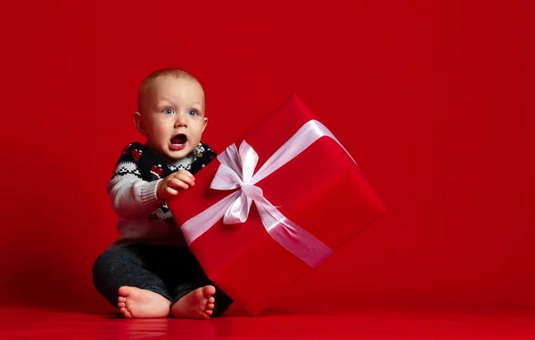 Хлопчик з великими блакитними очима в теплий светр, сидить перед його подарунком у загорнутій коробці зі стрічкою на червоному тлі . — стокове фото