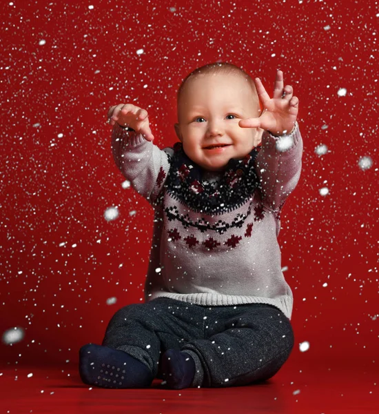 Lustiges kleines Baby im warmen gestrickten Weihnachtspullover im Studio an kalten Wintertagen. Es schneit Stockbild