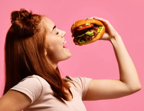 Крупным планом портрет голодной молодой женщины, поедающей бургер, изолированный на розовом фоне — стоковое фото