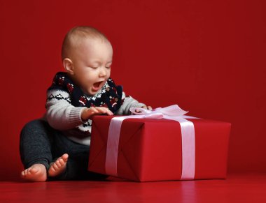 Mevcut onun önünde oturan sıcak kazak giyen iri mavi gözlü erkek bebek şerit kutu kırmızı arka plan üzerinde sarılı..