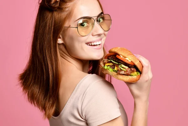 Εσωτερικη πορτρέτο του ένα πεινασμένο νεαρή γυναίκα τρώει burger που απομονώνονται σε ροζ φόντο — Φωτογραφία Αρχείου
