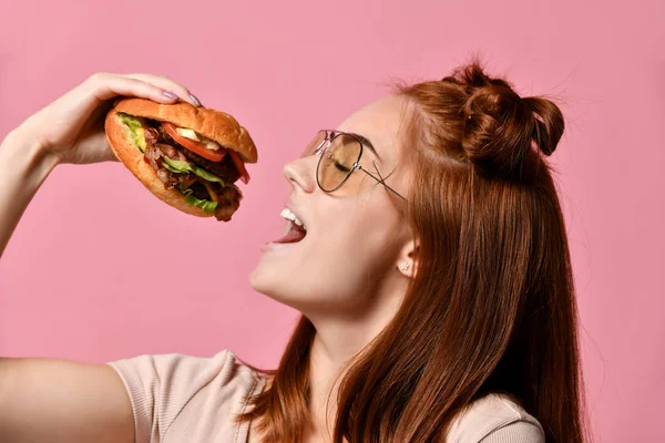 Närbild porträtt av en hungrig ung kvinna äta burger isolerade över rosa bakgrund — Stockfoto