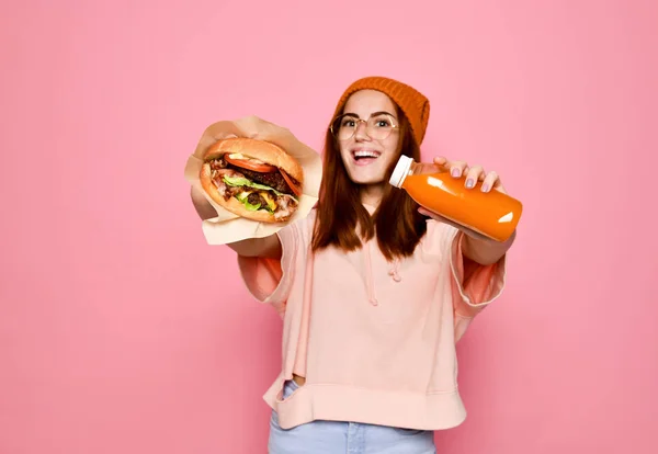 Vacker tonårsflicka med rött hår och hatt håller hamburgare och dryck i båda händerna. — Stockfoto