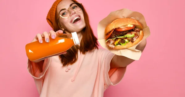 Kızıl saçlı ve şapka burger ve içecek her iki elinde tutan güzel genç kız. — Stok fotoğraf