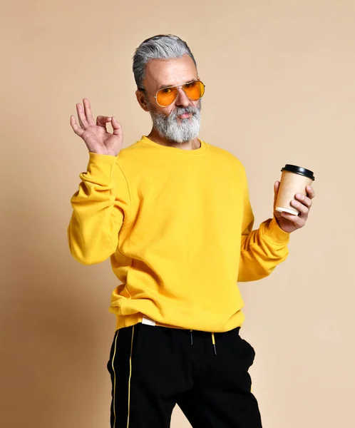 Hombre de moda senior mirando a la cámara, teniendo gorra con café en brazos y muestra un cartel bien con sus dedos — Foto de Stock