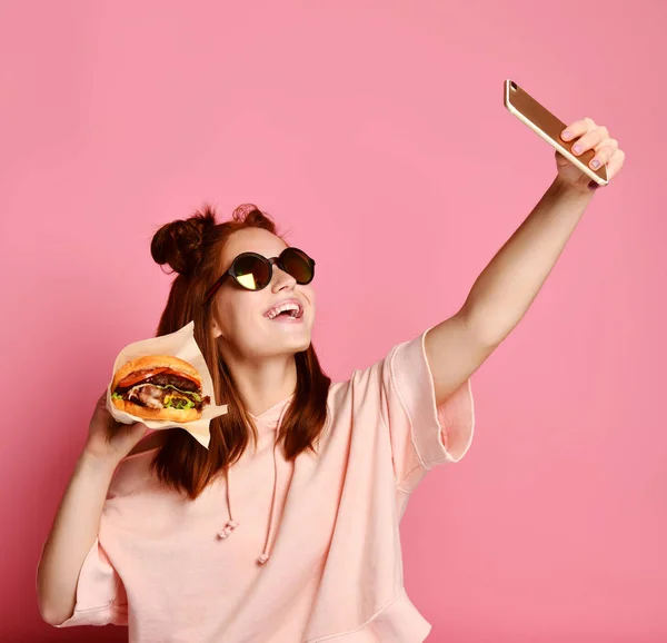 Chica sonriente en ropa de estilo de vida tomando foto autorretrato en el teléfono inteligente con sándwich de hamburguesa — Foto de Stock