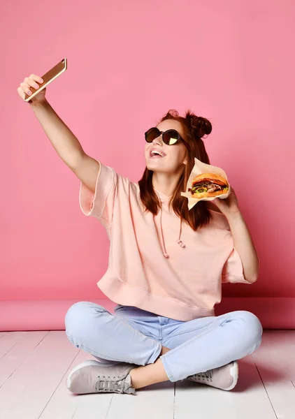 Alarak yaşam tarzı giysili kızı gülümseyen resim kendi kendine portre burger sandviç ile Smartphone — Stok fotoğraf