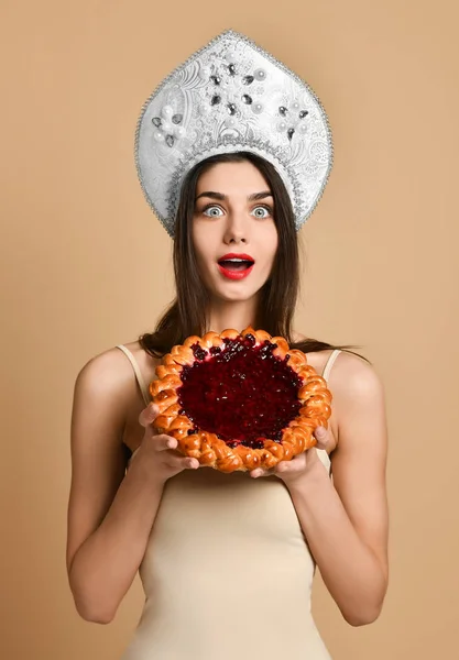Wanita Rusia yang cantik di topi kokoshnik, memiliki kue selama makan malam, terkejut dengan berdiri di atas latar belakang warna krem telanjang . — Stok Foto