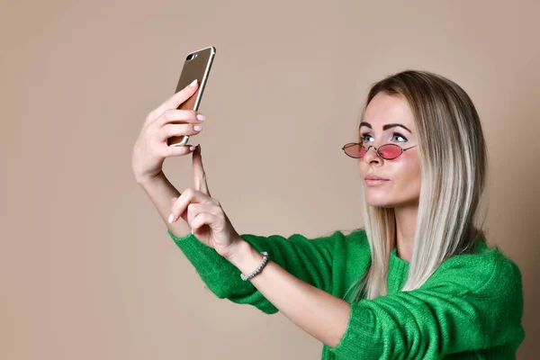 Retrato de cerca de la joven mujer rubia de moda alegre en ropa de jersey hace selfie en el teléfono inteligente, sobre fondo beige — Foto de Stock