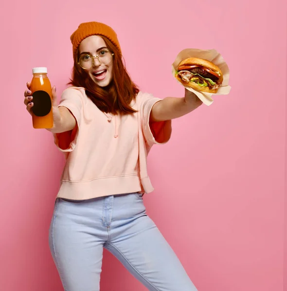 Vacker tonårsflicka med rött hår och hatt håller hamburgare och dryck i båda händerna. — Stockfoto