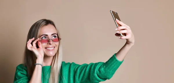 Retrato de cerca de la joven mujer rubia de moda alegre en ropa de jersey hace selfie en el teléfono inteligente, sobre fondo beige — Foto de Stock