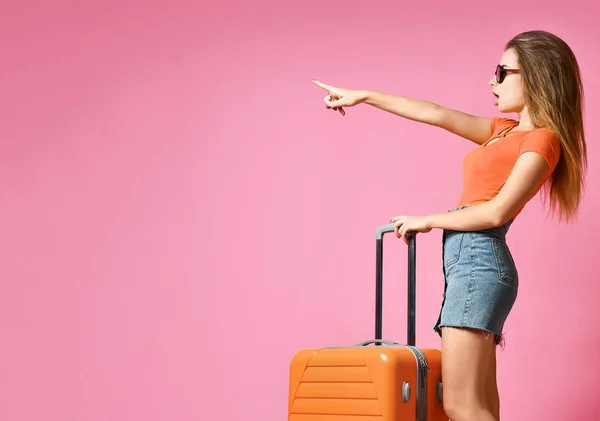 Portrait d'une jeune femme caucasienne joyeuse vêtue de vêtements d'été, avec une valise et pointant du doigt — Photo