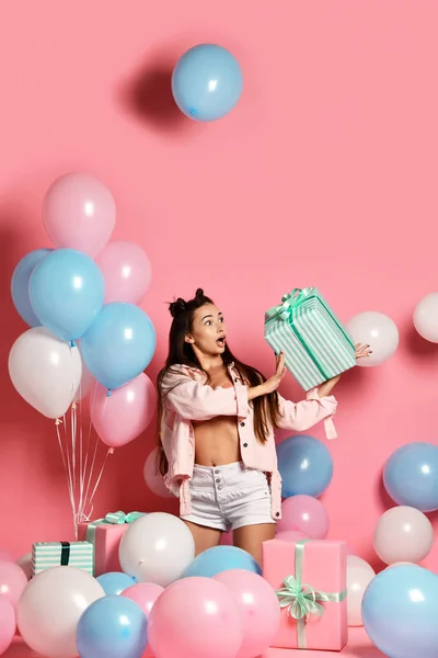 シャツとショーツ ギフトのカラフルな気球とパステル ピンクの背景に現在の箱を持ってかなりうれしそうな若い女性の肖像画。誕生日パーティ、人々 の心からの感情 — ストック写真