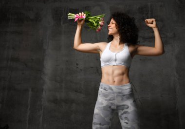 güzel vücut atletik kız kıvırcık saçlı ve spor giyim, ihale tulpan gülümseme bir buket güçlü bir kas kolundan tutar ve beton bir arka plan karşı tarafa görünüyor. Kadın güç