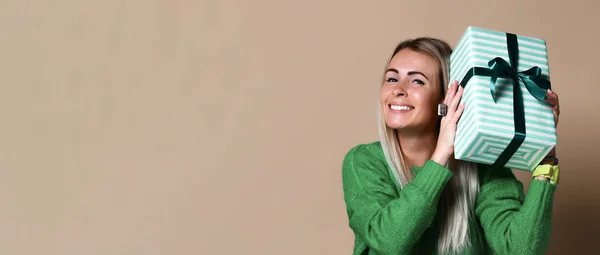 Retrato de una chica rubia bastante sonriente sosteniendo una caja de regalo y mirándola, aislada sobre un fondo beige de color —  Fotos de Stock