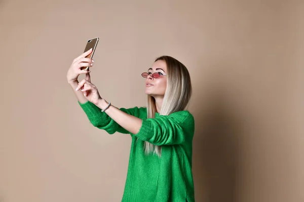 Retrato de cerca de la joven mujer rubia de moda alegre en ropa de jersey hace selfie en el teléfono inteligente, sobre fondo beige — Foto de Stock