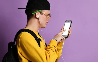 Şapkalı, gözlüklü ve mor arka planda akıllı telefon kullanan neşeli bir öğrencinin portresi.