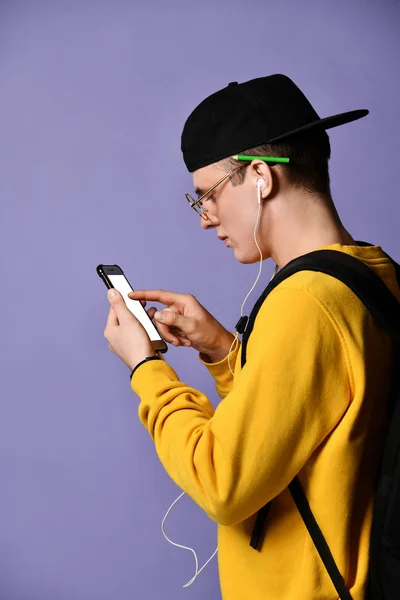 Portrait of a cheerful student wearing backpack, in cap and glasses and using smartphone over purple background — Stock Photo, Image