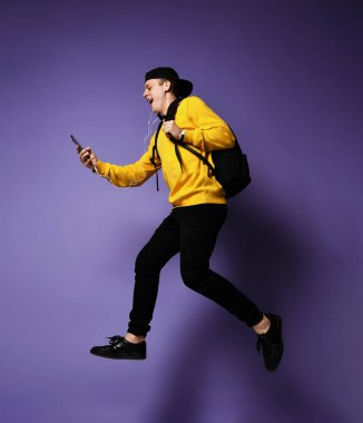 sırt çantası ve kulaklık ile rahat giyim öğrenci adam bir akıllı telefon kullanarak müzik dinliyor ve atlama sırasında gülümseyerek