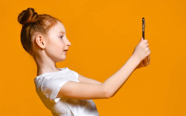 Alegre niña pelirroja tomando selfie con teléfono móvil contra una pared amarilla . — Foto de Stock