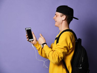 Şapkalı, gözlüklü ve mor arka planda akıllı telefon kullanan neşeli bir öğrencinin portresi.